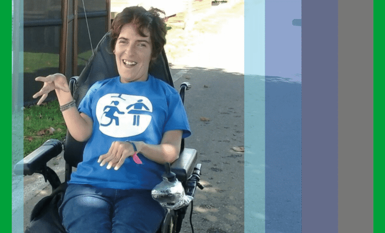 Marian Andres sentada en su silla eléctrica. Lleva una camiseta de la Asociación Cambiadores Inclusivos y está en un camino soleado con hojas.