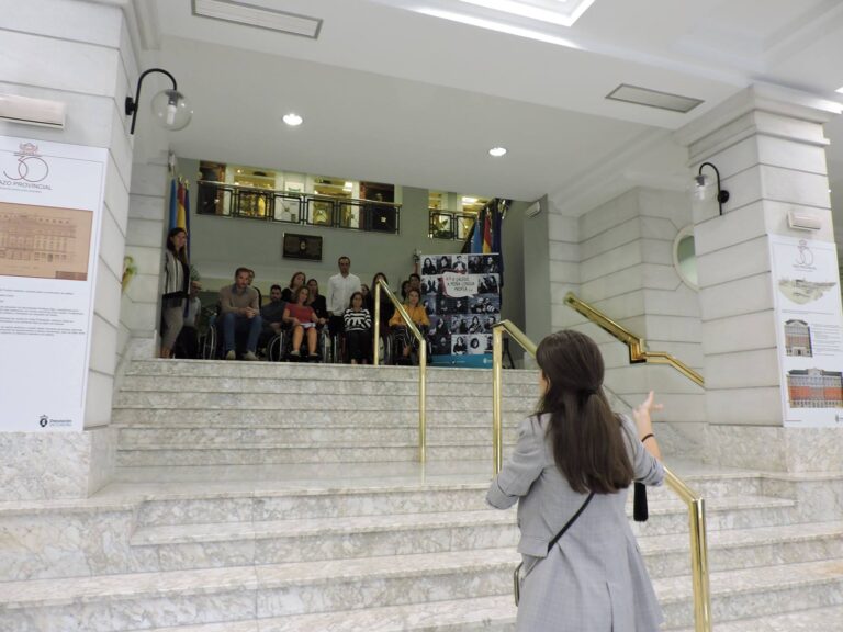 Foto de Belén Vaz. Esta de espaldas a la cámara situada al pie de unas escaleras. Arriba de las escaleras hay un pequeño grupo de personas a los que Belén se está dirigiendo.